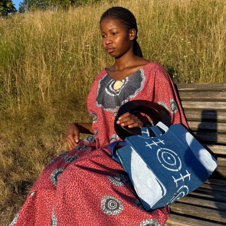 Robe Àkua en wax rouge à motifs cercles blancs portée par une femme assise sur un banc, avec sac Aabò en jean bogolan bleu - carre