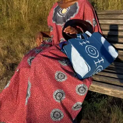 Robe Àkua en wax rouge à motifs cercles blancs, portée sur un banc avec le sac Aabò bleu