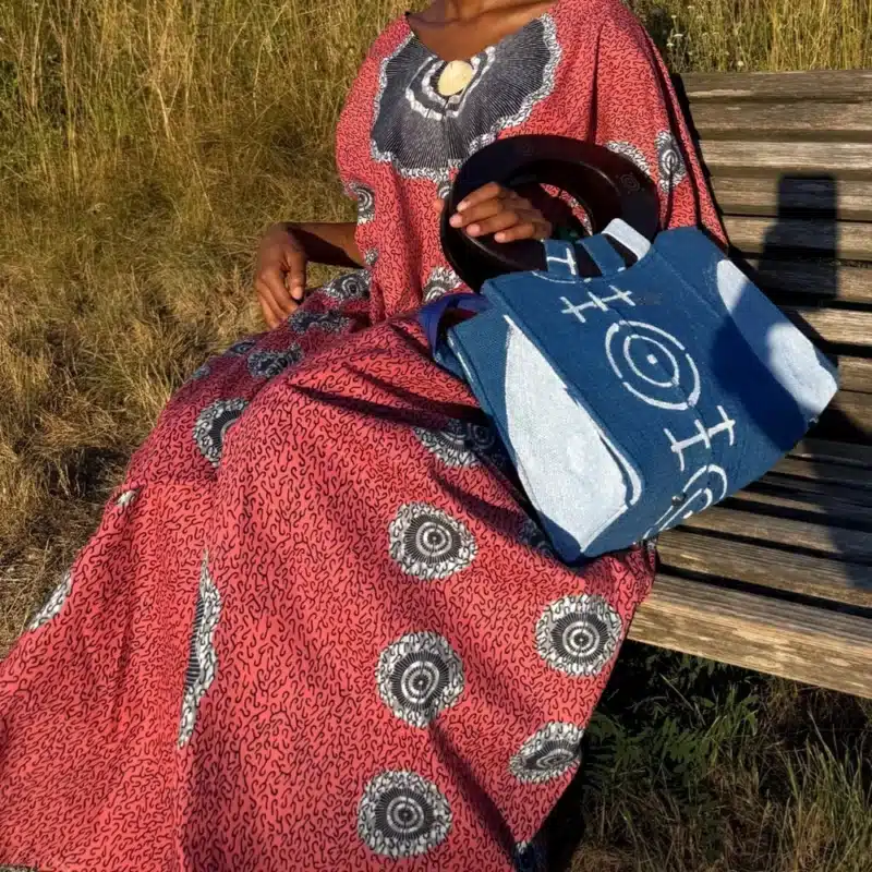 Robe Àkua en wax rouge à motifs cercles blancs, portée sur un banc avec le sac Aabò bleu