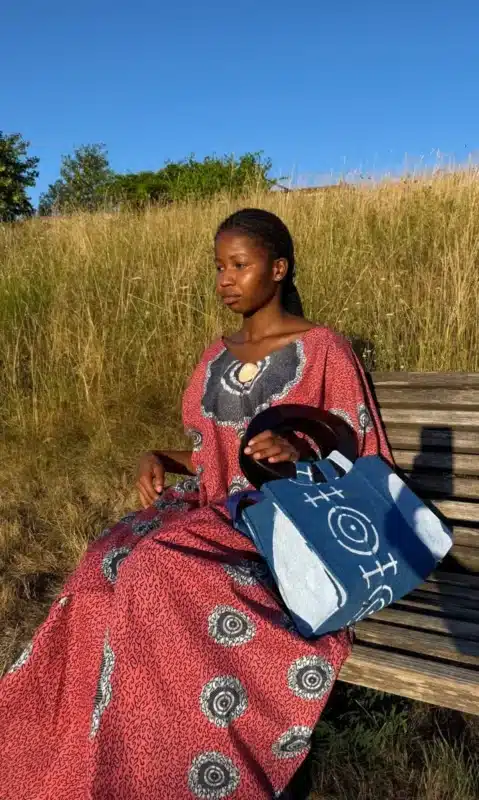 Robe Àkua en wax rouge à motifs cercles blancs portée par une femme assise sur un banc, avec sac Aabò en jean bogolan bleu