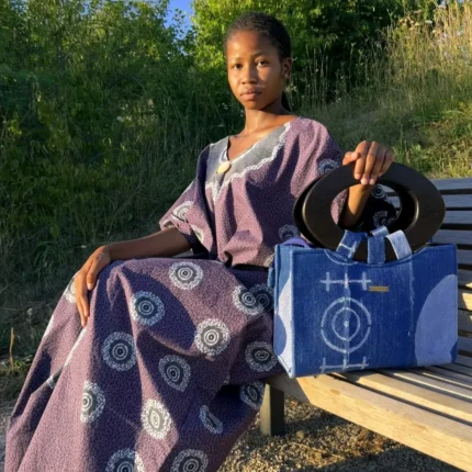 Robe Àkua en wax violet à motifs cercles blancs portée par une femme assise, avec sac Aabò en jean bogolan bleu
